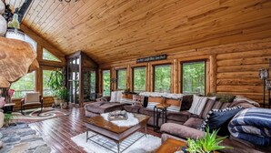 A cozy log cabin interior with a large wooden ceiling, a stone fireplace, a sofa set, and a dining table. - The Hideaway on Little Jay | Log Home | (Cohasset)