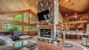 A spacious living area with a stone fireplace, wooden ceiling, and large windows. - The Hideaway on Little Jay | Log Home | (Cohasset)