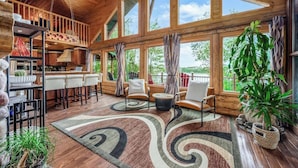 A spacious cabin with a wooden interior, large windows, a kitchen area, and a seating area with a view of the outdoors. - The Hideaway on Little Jay | Log Home | (Cohasset)