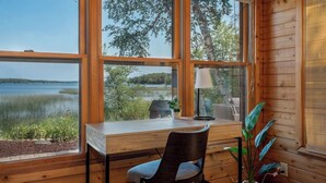 A cabin with a wooden interior, a desk, a chair, and a view of a lake and trees.