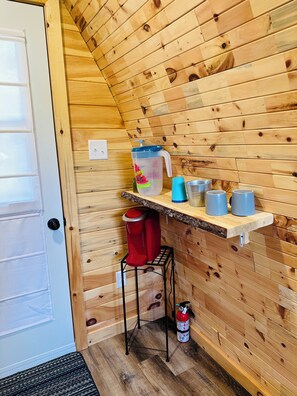 A small kitchenette with wooden walls, a coffee maker, a water jug, and a fire extinguisher.
