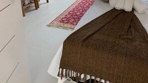 Un dormitorio con cama, televisión montada en la pared, una silla, una mesita y una ventana con cortinas.