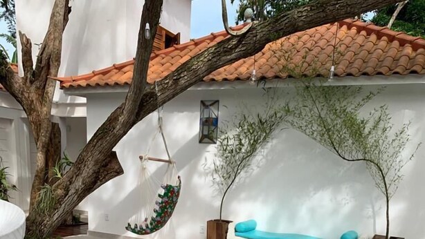 Una piscina con borde de baldosas a cuadros, pared blanca, techo de tejas rojas, una silla colgante y un banco con cojín azul.
