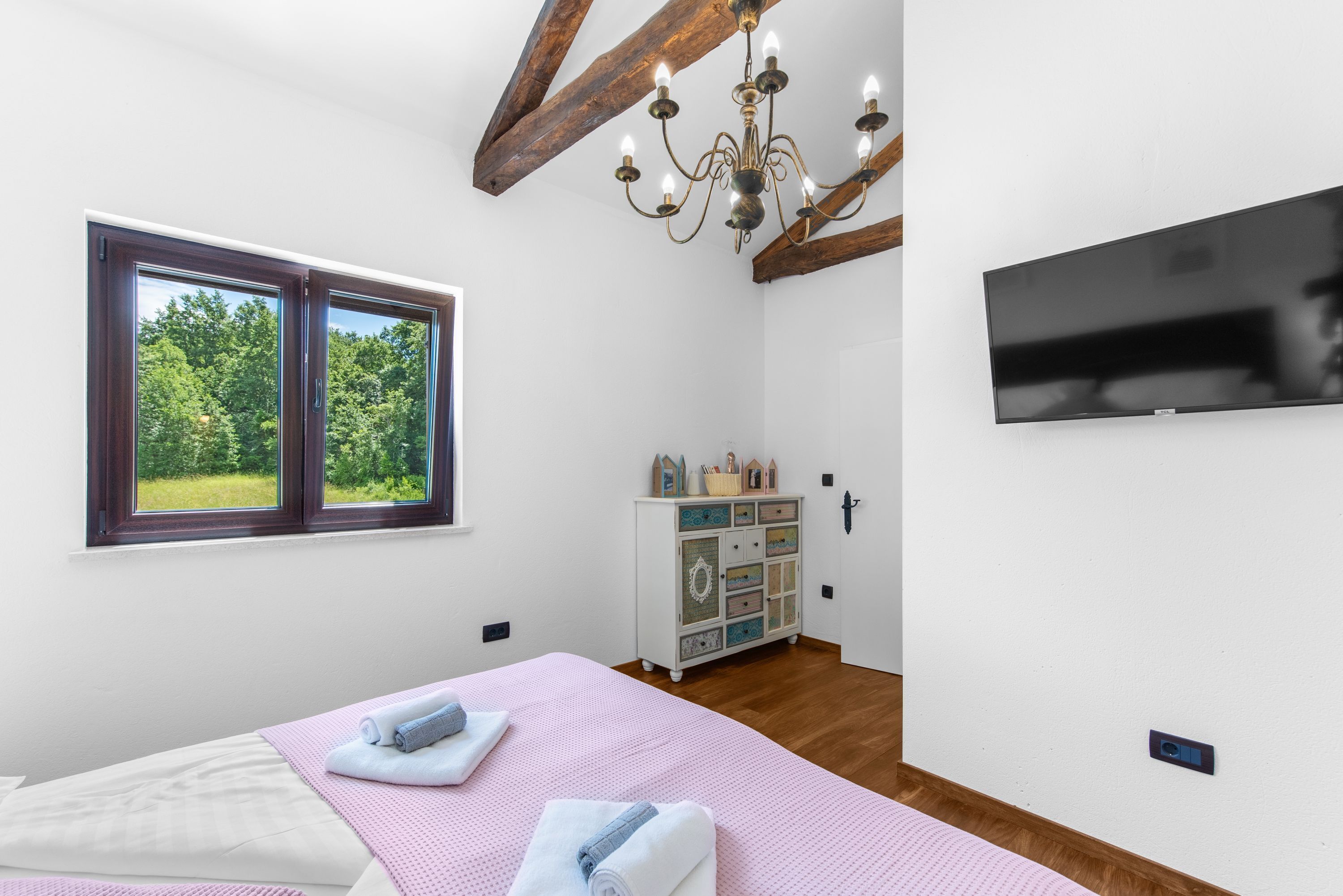 A bedroom with a bed, a chandelier, a television, a dresser, and a window with a view of greenery.
