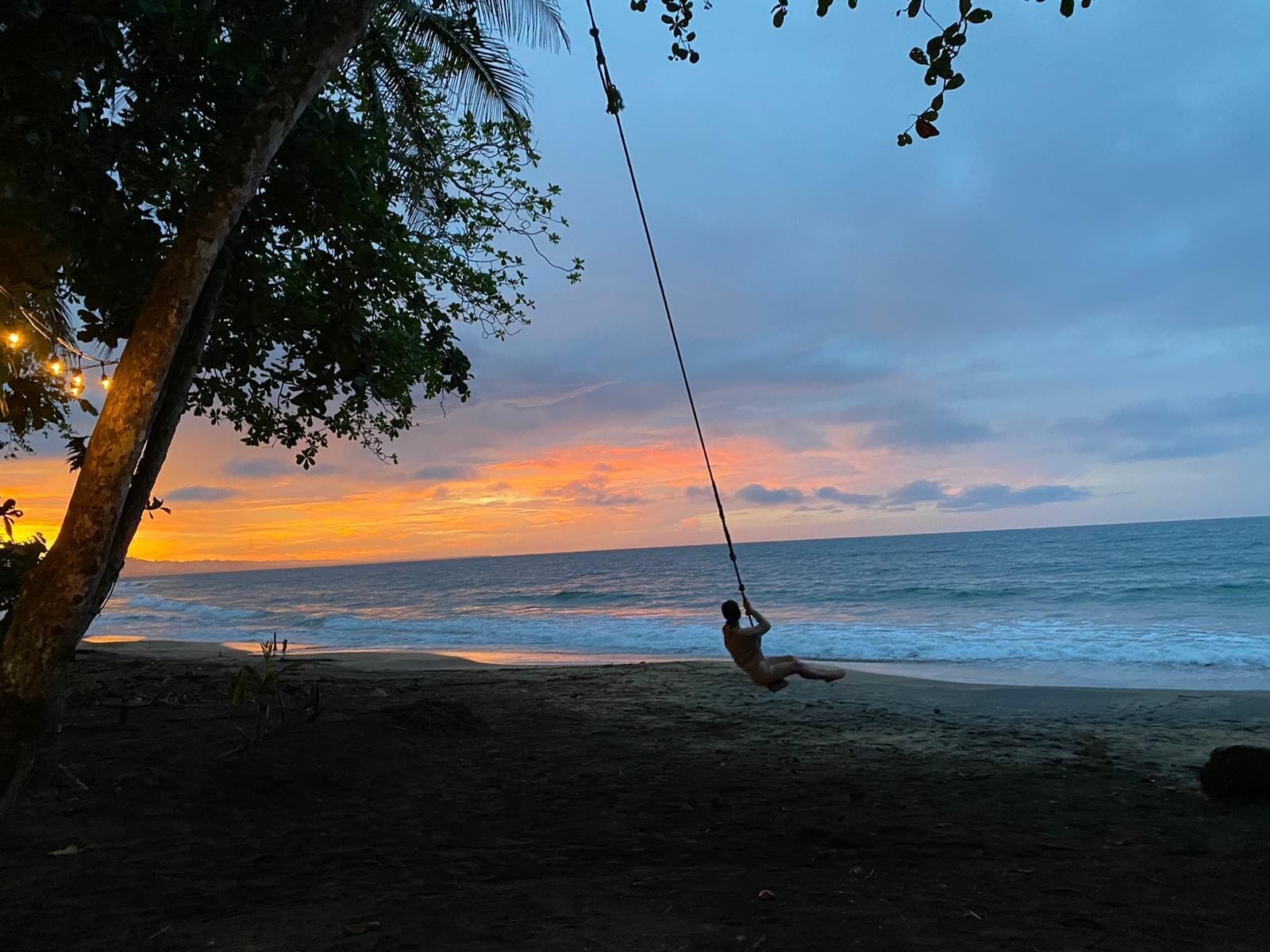 A beach scene with a person on a swing overlooking the ocean at sunset.