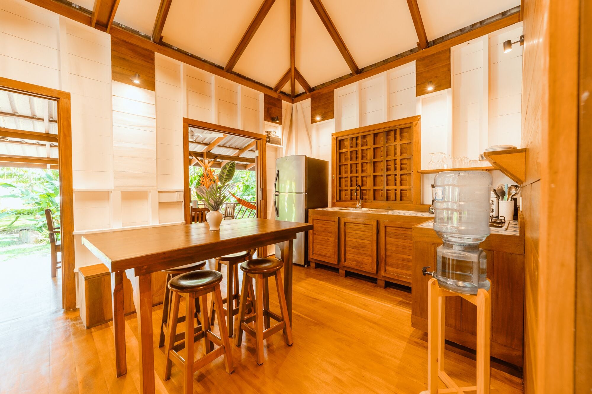 Uma cozinha com mesa de madeira, banquetas, geladeira e bebedouro.
