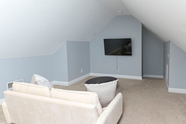 Une pièce avec un plafond haut, un téléviseur à écran plat fixé au mur, un canapé beige et un pouf rond.