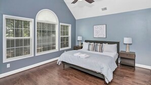 A bedroom with a bed, two bedside tables, a ceiling fan, and large windows with blinds.