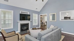 A living room with a fireplace, a television, a ceiling fan, and a kitchen area visible through an open door.