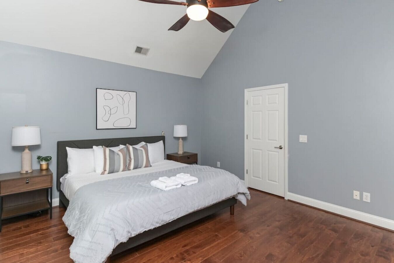 A bedroom with a bed, two bedside tables, a ceiling fan, a door, and a wall-mounted artwork.