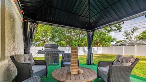Eine überdachte Terrasse mit Korbmöbeln, einem Holz-Jenga-Turm und einem Grill.