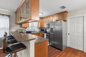 Uma cozinha com armários de madeira, uma geladeira de aço inoxidável, micro-ondas e forno embutido. Há banquetas e uma grande ilha com bancada de granito.