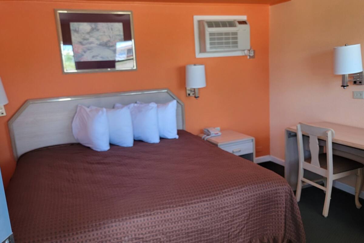A hotel room with a bed, a desk, a chair, a nightstand, a lamp, and an air conditioning unit.