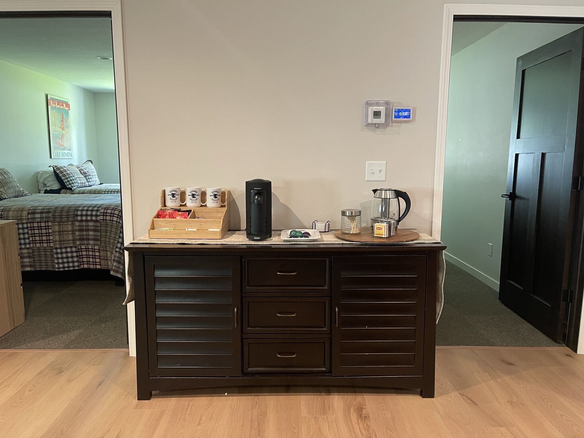 A dark wooden desk with drawers, a coffee maker, and a kettle. There is a bed with a plaid comforter and pillows in the background.
