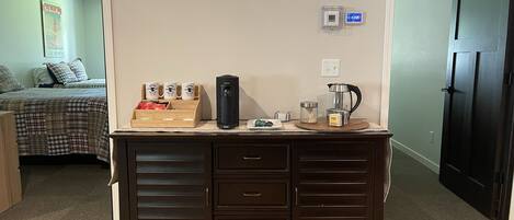 A dark wooden desk with drawers, a coffee maker, and a kettle. There is a bed with a plaid comforter and pillows in the background.