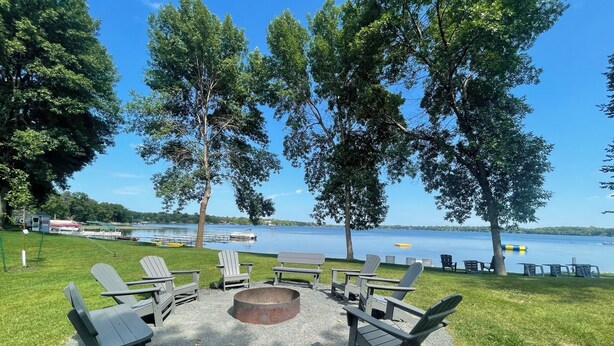 A lakeside outdoor seating area with Adirondack chairs and a fire pit, surrounded by trees and a view of the lake. - Guest House at Geneva Beach Resort - Views of Lake Geneva (Alexandria)