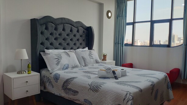 A bedroom with a tufted headboard, a bed with floral bedding, a bedside table with a lamp and a bottle, and a view of the city through large windows. - Zona céntrica Lima Histórica (Lima)