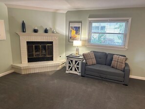 A living room with a fireplace, a gray sofa, a side table with a lamp, and a window with blinds.