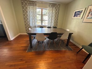 Un coin repas avec une table en bois, quatre chaises, des rideaux à motifs et deux tableaux encadrés accrochés au mur.