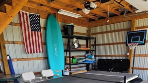 Une table de ping-pong dans un garage, avec un drapeau américain, une planche de surf et un panier de basket.