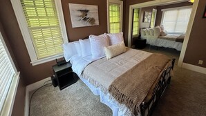 A bedroom with a large bed, white bedding, a bedside table with a lamp, and a painting on the wall. - Stay in Our Quaint Cottage Retreat for a Memorable Escape! (Duncan)