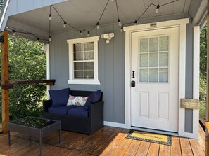 A porch with a white door, a bench with cushions, a glass table, and string lights.
