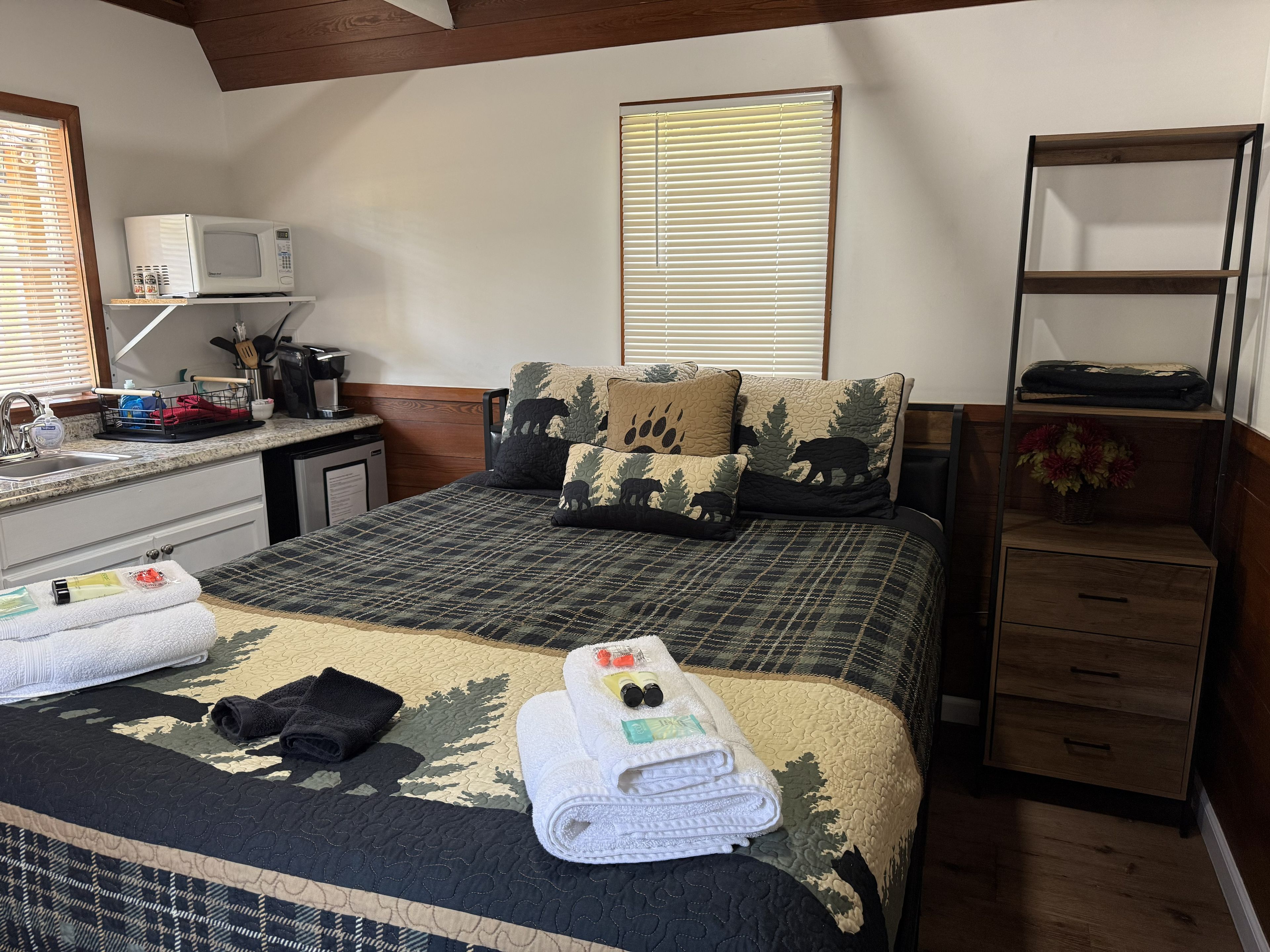 Un dormitorio con una cama, una mesita de noche, una lámpara, un ventanal con persianas, una estantería con objetos y suelo de madera.