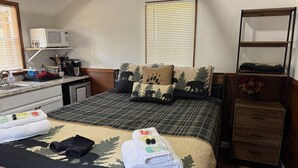 A bedroom with a bed, a bedside table, a lamp, a window with blinds, a shelf with items, and a wooden floor. - Casita 1, Tiny Home in Wood Area, 1 Mile From Glacier Entrance- 2 Guests (West Glacier)