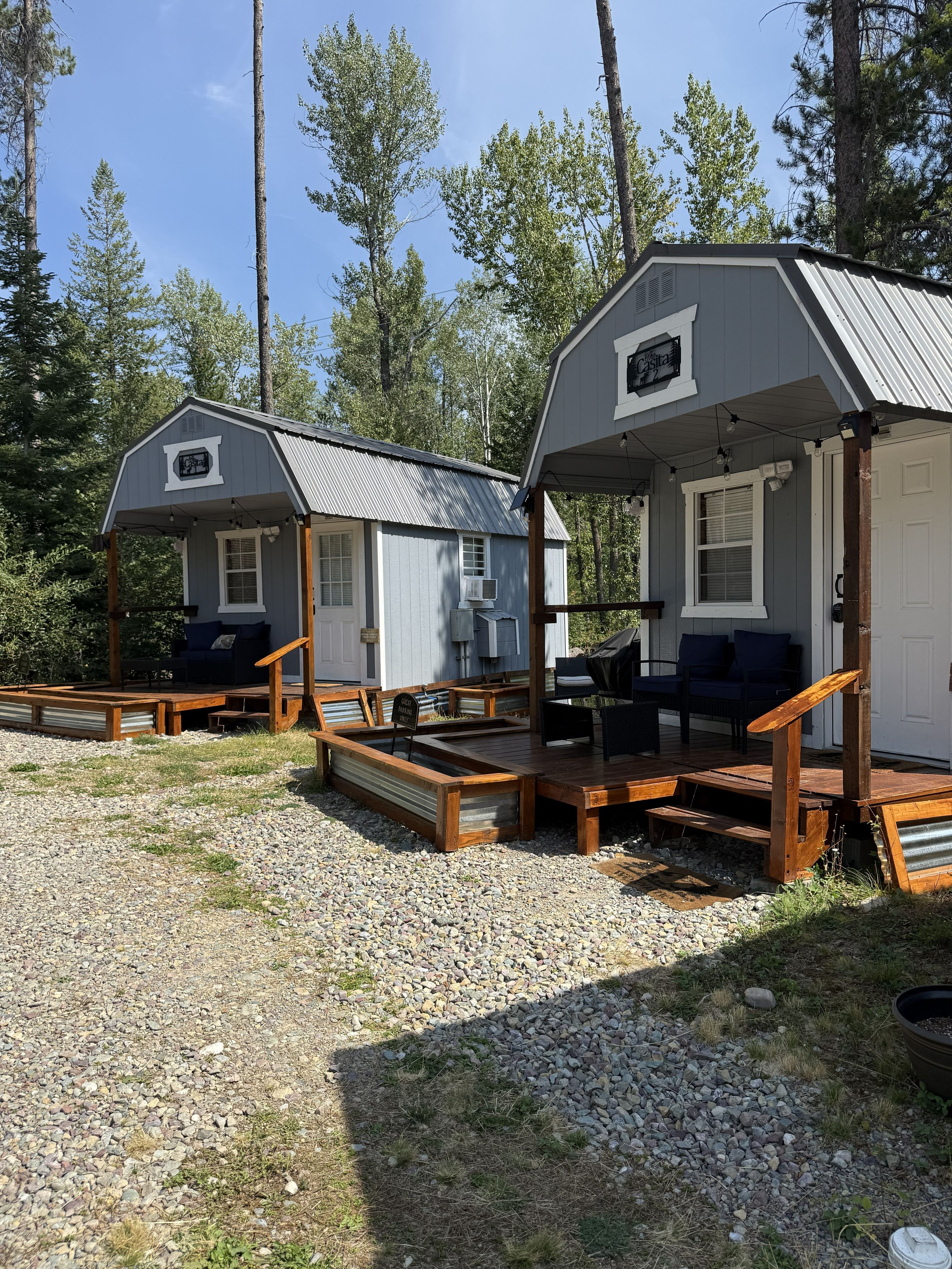 Dos cabañas con terrazas de madera y mobiliario exterior, rodeadas de árboles.