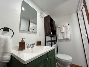 Un baño con lavamanos blanco, mueble de baño verde y un inodoro blanco. Hay un espejo sobre el lavamanos y un perchero con toallas.