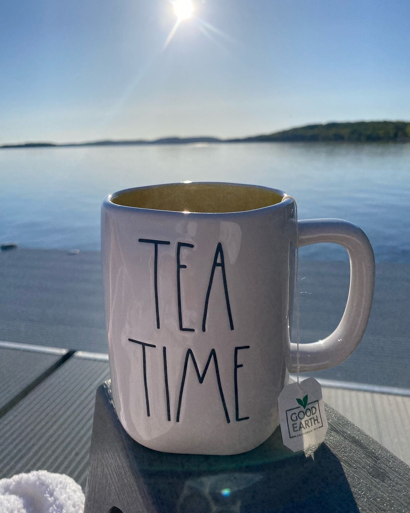 A white mug with "TEA TIME" written on it, placed on a table with a view of a calm body of water and a clear sky in the background.