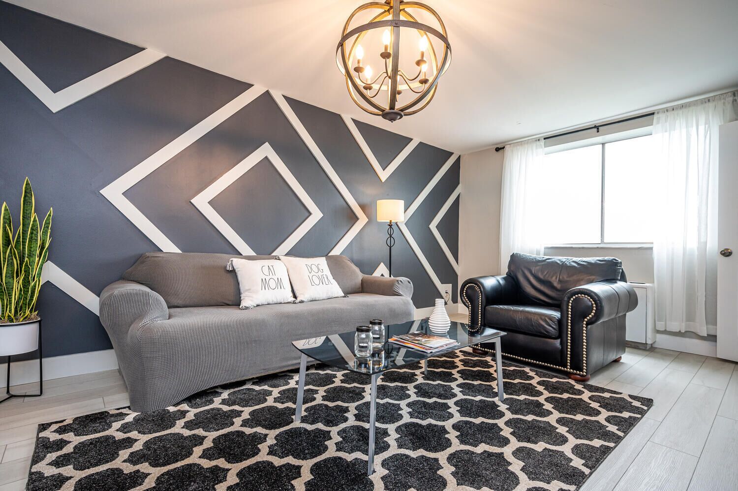 Un salon moderne avec un canapé gris, un fauteuil en cuir noir, un tapis à motifs et un motif géométrique sur le mur.