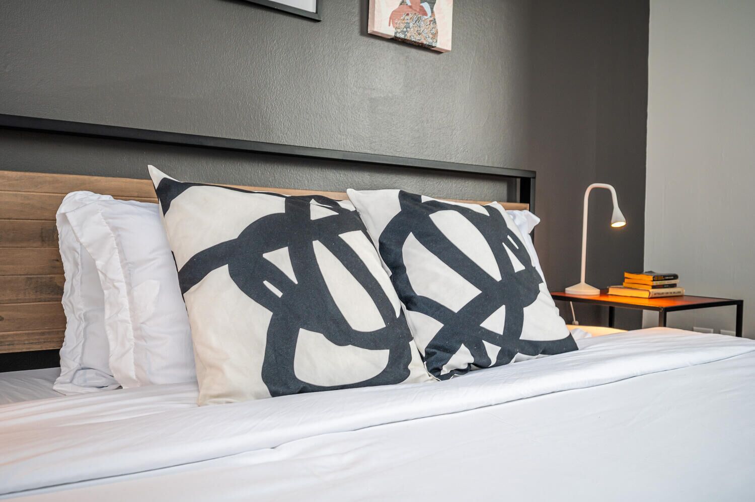 Un lit bien fait, avec une literie blanche, deux coussins noirs et blancs à motifs abstraits, une tête de lit en bois, une petite table de chevet avec une lampe et des livres, et un tableau encadré accroché au mur.