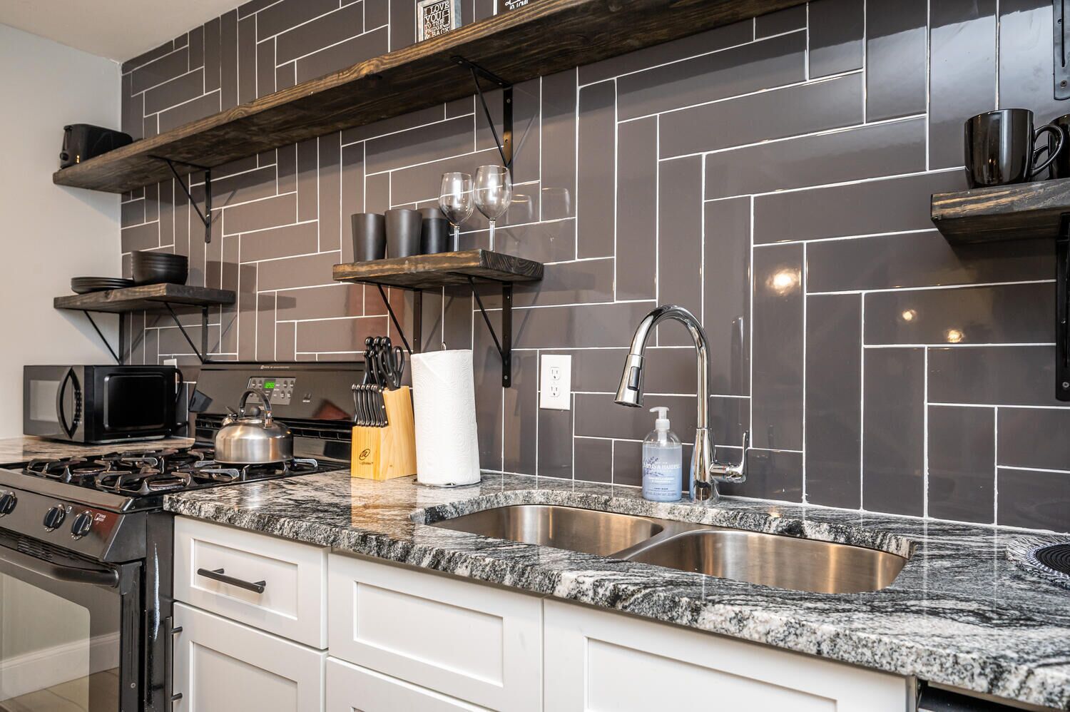 A kitchen with a granite countertop, stainless steel sink, and a gas stove. There are wooden shelves above the counter holding kitchenware and a kettle.