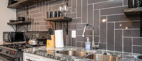 A kitchen with a granite countertop, stainless steel sink, and a gas stove. There are wooden shelves above the counter holding kitchenware and a kettle.