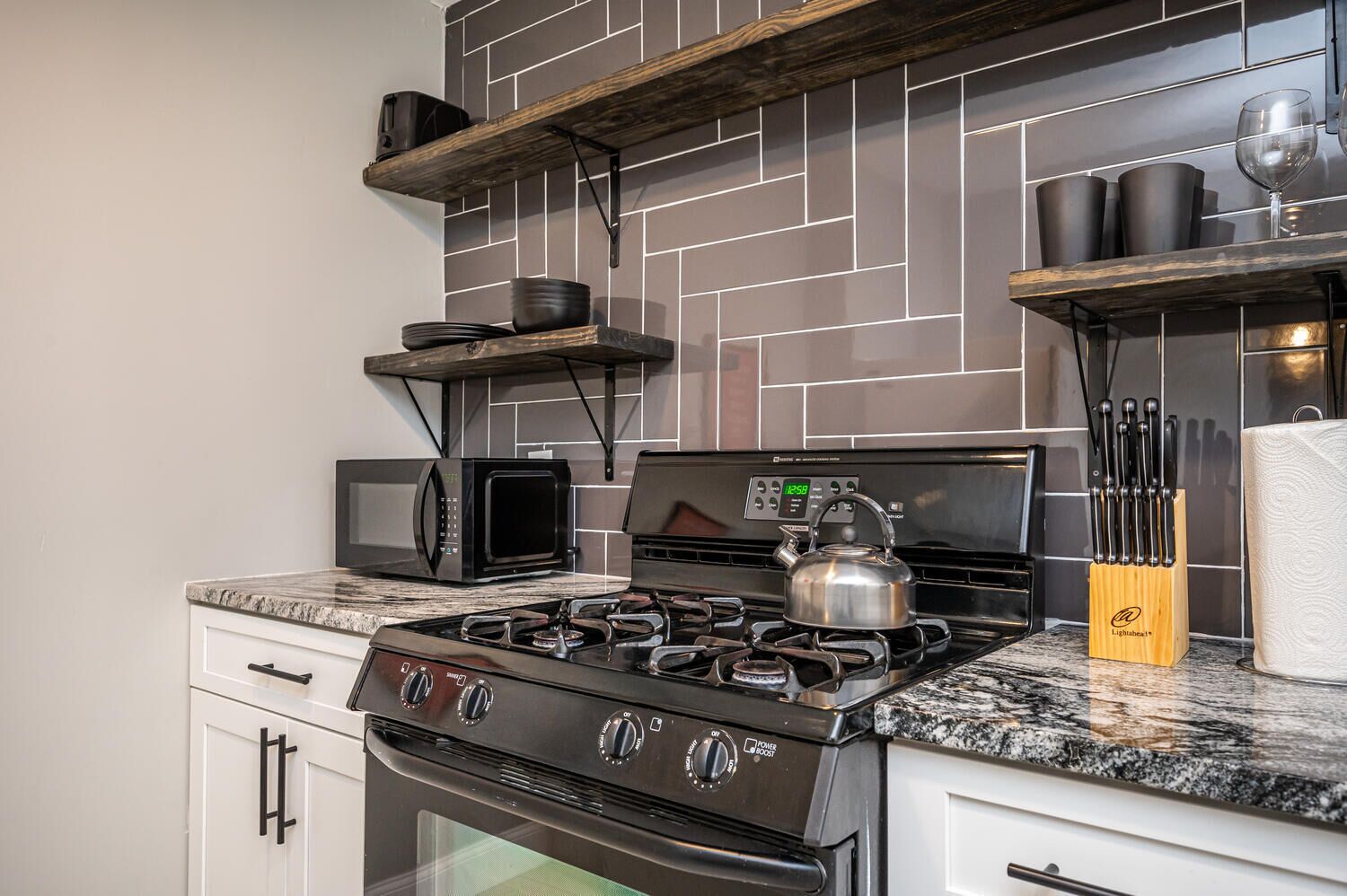 A kitchen with a gas stove, microwave, kettle, and various utensils on shelves.