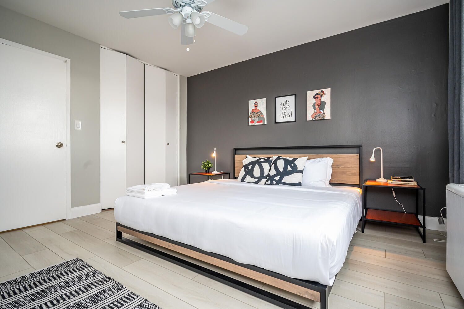 A modern bedroom with a large bed, a ceiling fan, a bedside table with a lamp, and artwork on the wall.