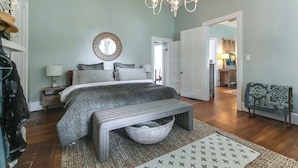 A bedroom with a bed, bedside tables, a chandelier, a mirror, and a view into another room. - 1883 Colonial Revival Historical Renovation  (Mount Pleasant)