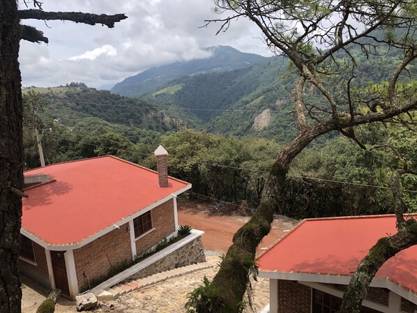 Un edificio con techo rojo, paredes de ladrillo y base de piedra. Está rodeado de abundante vegetación y montañas al fondo.