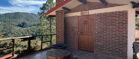 Una terraza de madera con una mesa y sillas, un edificio de ladrillo con techo rojo y vistas a una colina boscosa.