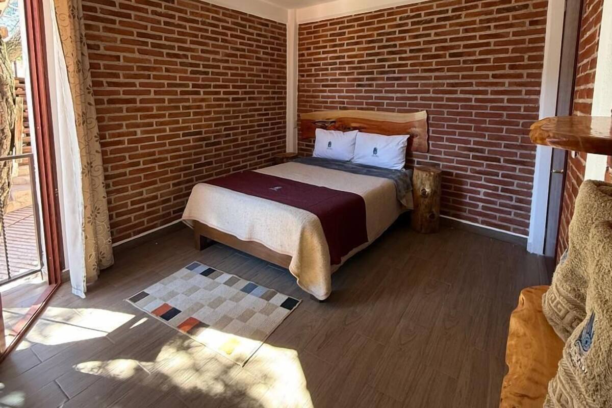 A bedroom with a brick wall, a bed with a wooden headboard, a wooden floor, and a window with curtains.