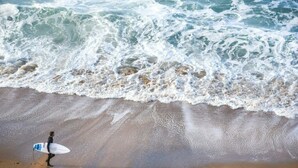 Una persona porta un surfboard su una spiaggia sabbiosa, con le onde che si infrangono sullo sfondo.
