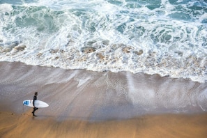 Una persona lleva una tabla de surf en una playa arenosa, con olas rompiendo al fondo.