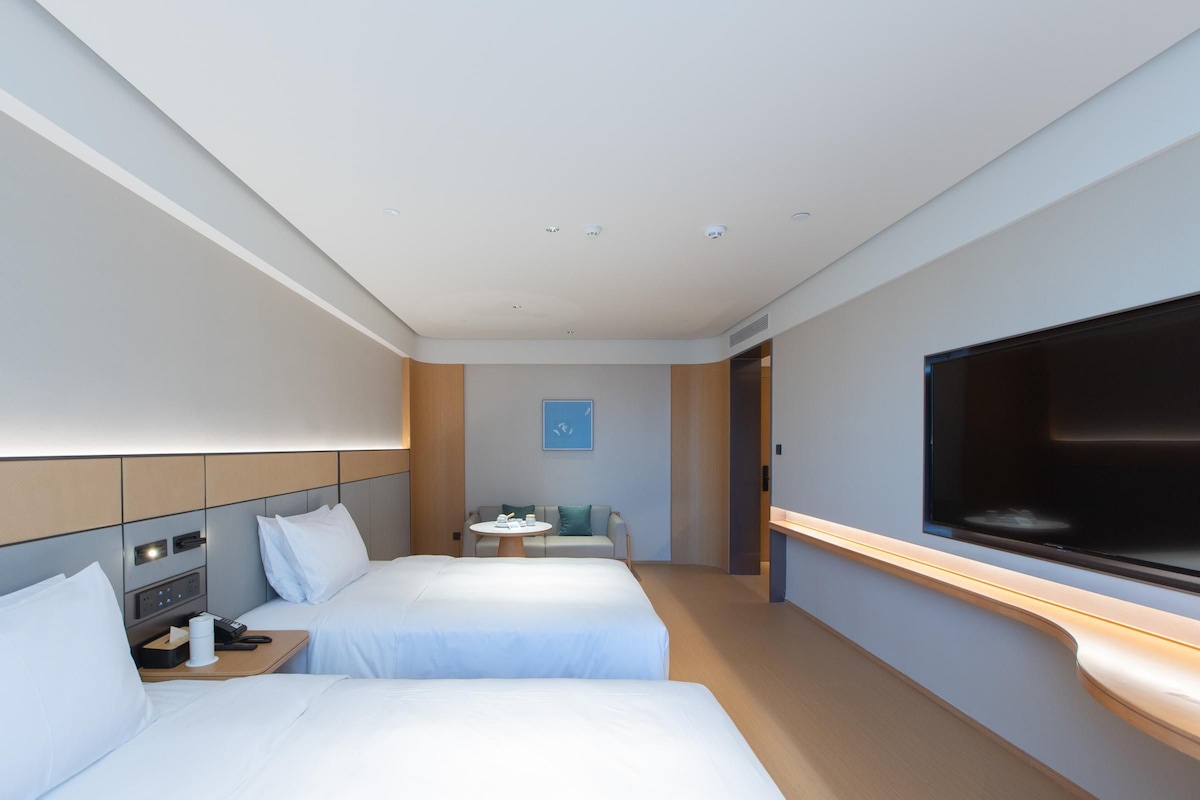 A hotel room with two beds, a TV, a small table with a chair, and a wall-mounted TV.