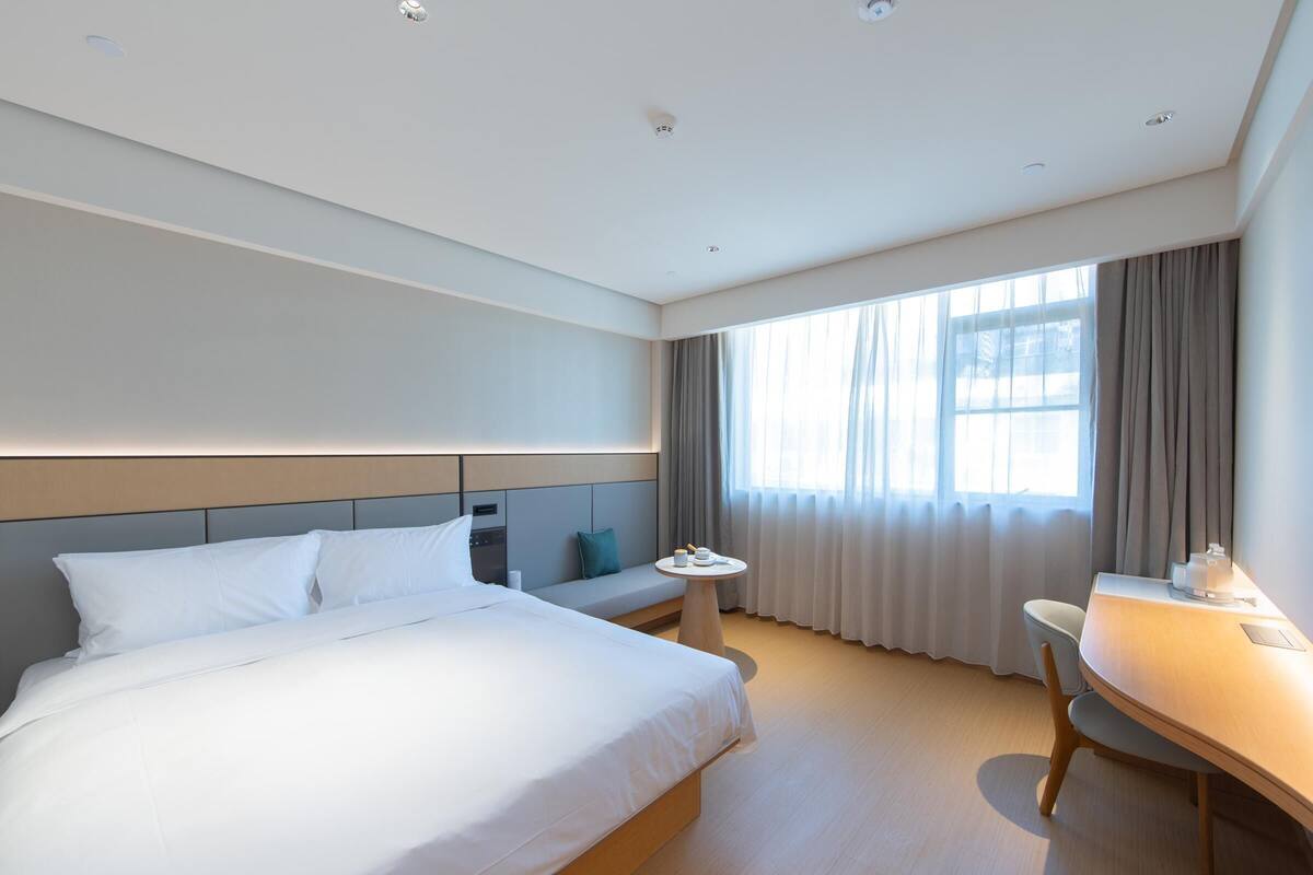 A modern hotel room with a large bed, a small round table, a desk, and a window with curtains.