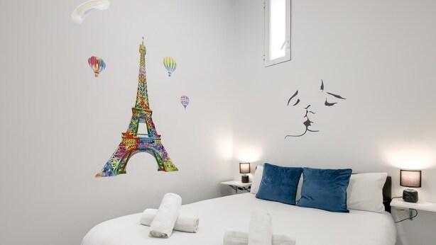 Un letto rifatto con cuscini bianchi e blu, una coppia di asciugamani arrotolati, e una parete con la Tour Eiffel e sagome di uccelli colorati.