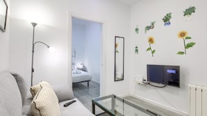 Un soggiorno moderno con un divano grigio, un tavolino in vetro, una TV a schermo piatto montata a parete e un corridoio che conduce a una camera da letto.