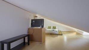 Quarto pequeno e moderno com piso de madeira, uma cama com almofadas verdes, uma televisão em um suporte e um banco.