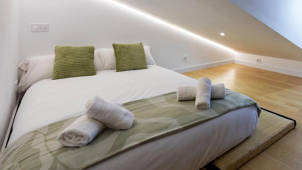 Quarto moderno com teto inclinado, cama com roupa de cama branca e almofadas verdes, e piso de madeira.