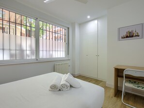 A hotel room with a large bed, a radiator, a window with blinds, a wardrobe, and a desk with a chair.